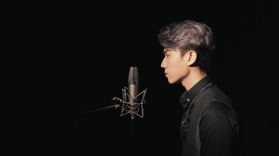 A young Asian male singer recording in a studio with a condenser microphone against a black background.